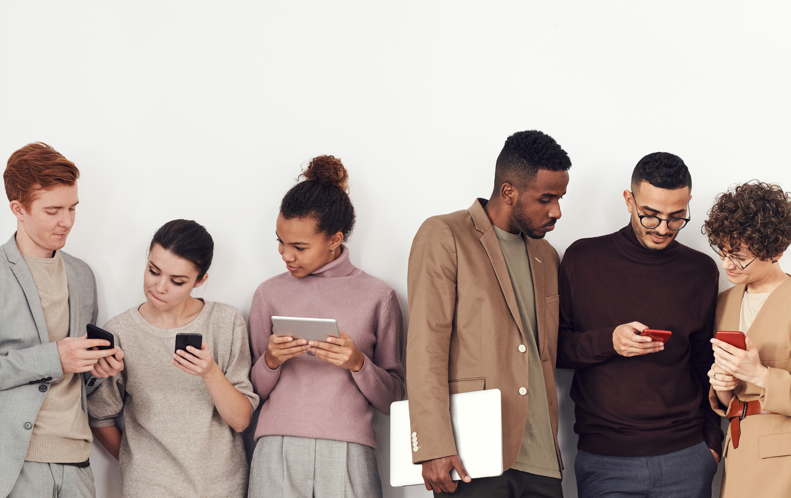 The pictures shows a group of six people of different genders and ethnicities in front of a white wall dressed in smart casual. They are each holding a phone, tablet or laptop.