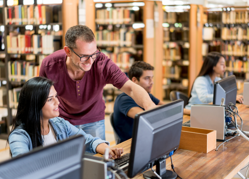 The picture shows a library. In the foreground there is a long desk with three stationary computers. On the nearest computer a male teachers explains something to a female student whilst pointing to the screen.